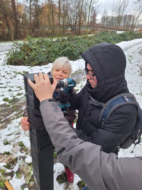 Twee mensen bevestigen een bord in een besneeuwde, bosrijke omgeving.