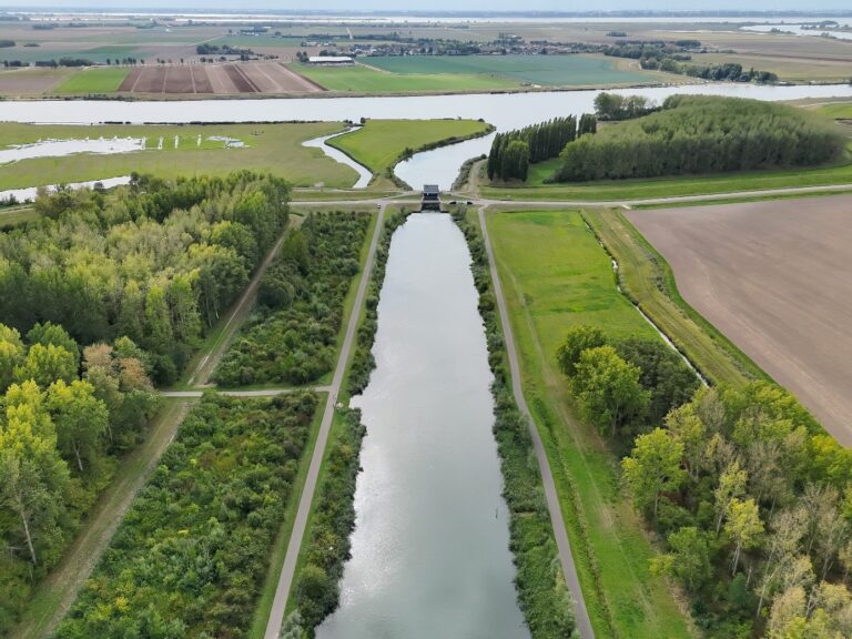 Kanaal tussen groene bossen, met brug en landbouwvelden in de verte.