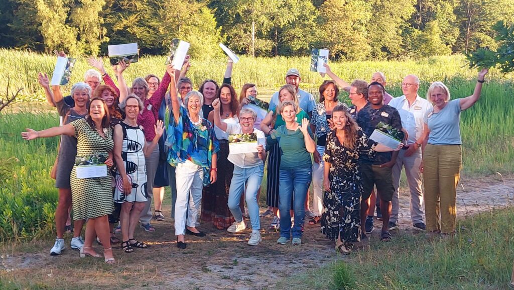 Groep mensen viert iets in een groene, zonnige buitenomgeving met opgeheven armen en papieren.