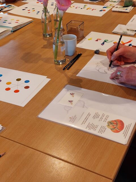 Een persoon schetst op papier aan een tafel met kleurstalen en bloemen.