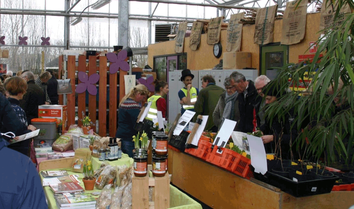 Een drukbezochte markt in een kas met planten en producten uitgestald op tafels. Mensen bekijken goederen.