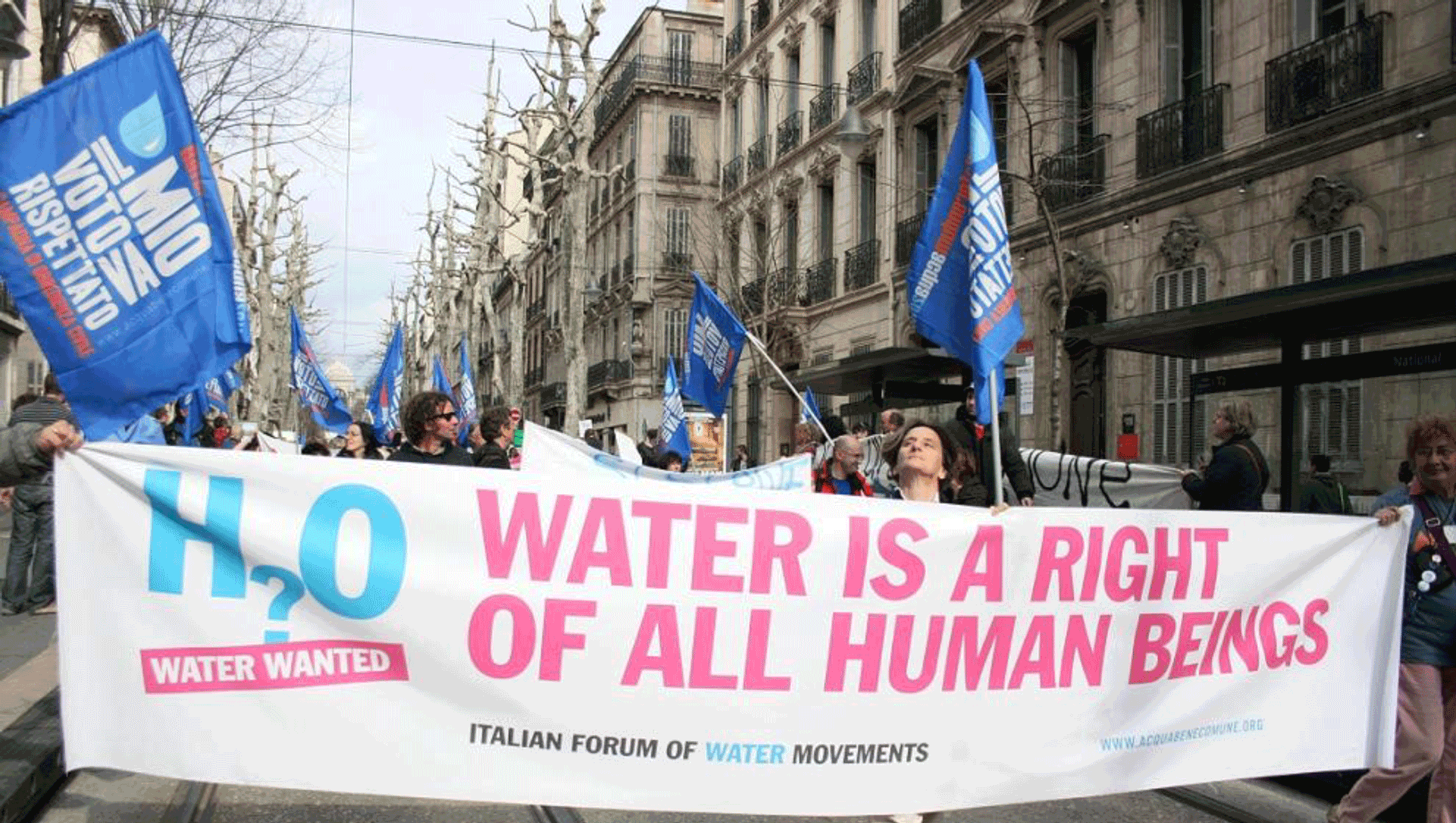 Demonstranten met spandoek "Water is a right of all human beings", blauwe vlaggen in stadsstraat.