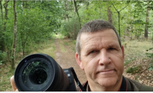 Man met camera in een bosgebied, met een bospad en groene bomen op de achtergrond.