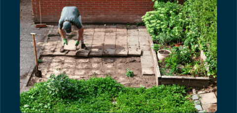 Persoon legt tegels in de tuin naast een verhoogd bloembed met groene planten.