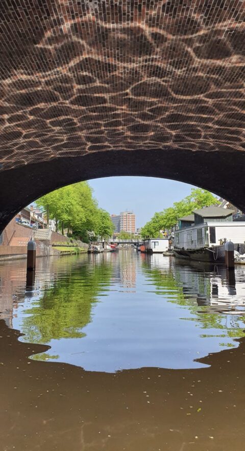 Kanaal onder een bakstenen brug met bomen en gebouwen op de achtergrond.