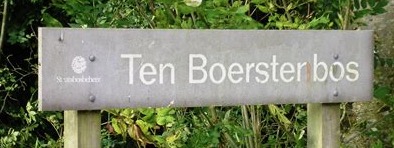 Bord met tekst "Ten Boersterbos" omgeven door groene struiken.