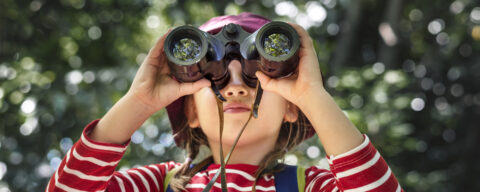 Kind met een rode hoed en streepjestrui kijkt door een verrekijker in een bosachtige omgeving.